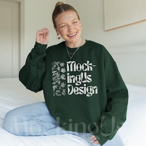 Op de afbeelding: Een donker bosgroene sweater met ronde hals en een witte grafische print op de borst. Het ontwerp bevat een verticale schikking van lijntekeningen van bloemen aan de linkerkant en de tekst Mock-ingUs Design in een golvend lettertype.