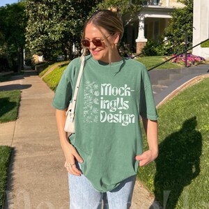 Op de afbeelding: Een saliegroen T-shirt met korte mouwen, voorzien van een witte bloemenafbeelding aan de linkerkant en de tekst Mock-ingUs Design in een gestileerd wit lettertype. Het shirt heeft een ronde hals en een relaxte pasvorm, gecombineerd met een lichtblauwe spijkerbroek.