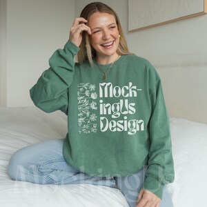 May include: A green sweatshirt with the text "Mockings Design" in white. The sweatshirt has a floral design on the left side. The person is wearing blue jeans and sitting on a white bed.