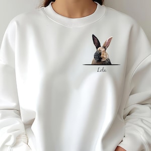 May include: White sweatshirt with a brown and black rabbit peeking out of a pocket on the front. The text "Lola" is printed below the rabbit.