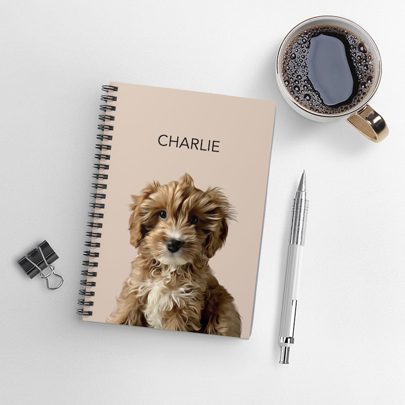 May include: A spiral-bound notebook with a light beige cover featuring a digital illustration of a fluffy brown and white puppy and the name CHARLIE printed in black text. The notebook is positioned next to a silver pen and a black binder clip.