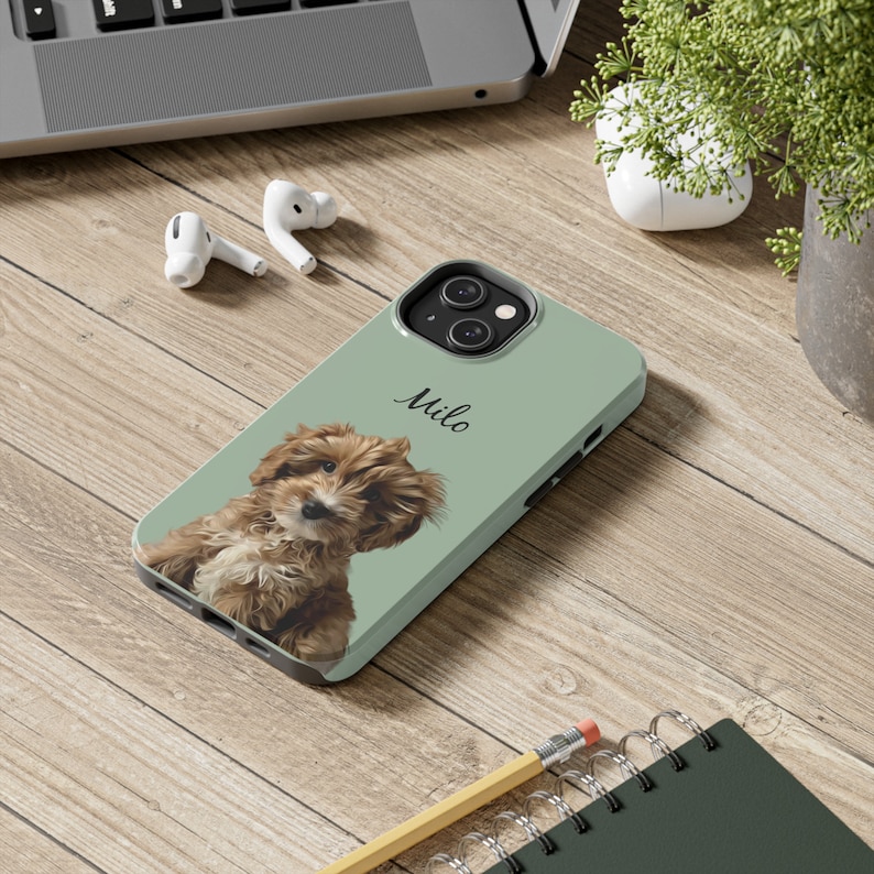 May include: A light green phone case featuring a brown and white puppy with the name "Milo" in a handwritten font. The case is on a wooden surface with a laptop, earbuds, and a small plant in the background.
