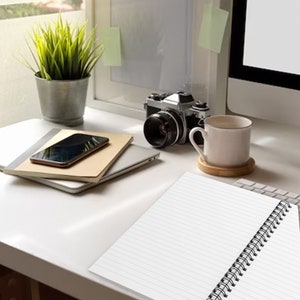 May include: A white spiral-bound notebook with lined pages sits on a desk alongside a smartphone, a silver laptop, a vintage-style camera, a potted green plant in a metallic container, and a white ceramic mug on a wooden coaster.