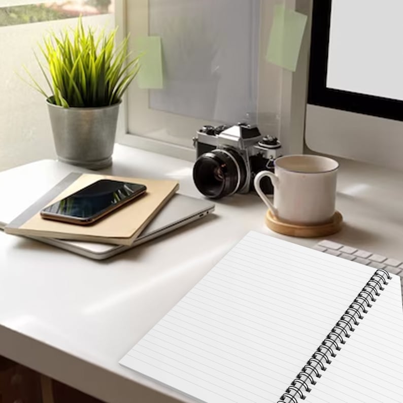 May include: A white spiral-bound notebook with lined pages sits on a desk alongside a smartphone, a silver laptop, a vintage-style camera, a potted green plant in a metallic container, and a white ceramic mug on a wooden coaster.