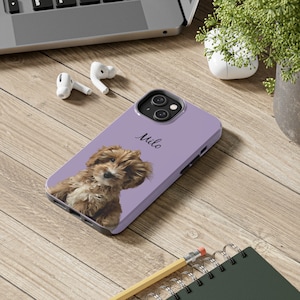 May include: A lavender phone case featuring a brown and white puppy with the name "Milo" in script. The case is on a wooden surface with a laptop, earbuds, and a pencil.