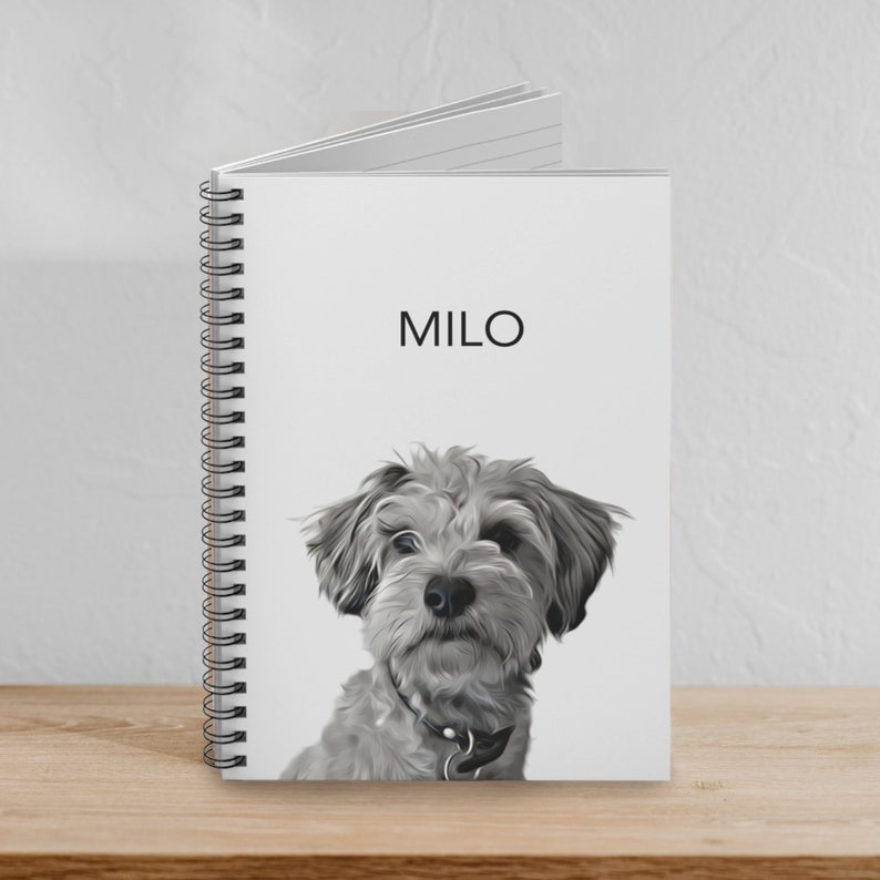 May include: A spiral-bound notebook with a white cover featuring a black and white illustration of a shaggy dog and the name MILO printed in black capital letters above the dog. The notebook has a black metal coil spine on the left side.