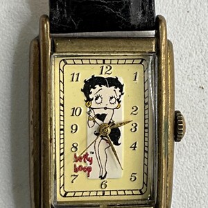Peut inclure: Une montre-bracelet rectangulaire avec un bracelet en cuir noir et un bo&icirc;tier dor&eacute;. Le cadran est de couleur cr&egrave;me et repr&eacute;sente le personnage de dessin anim&eacute; Betty Boop. Les aiguilles sont dor&eacute;es et la montre a une petite couronne.