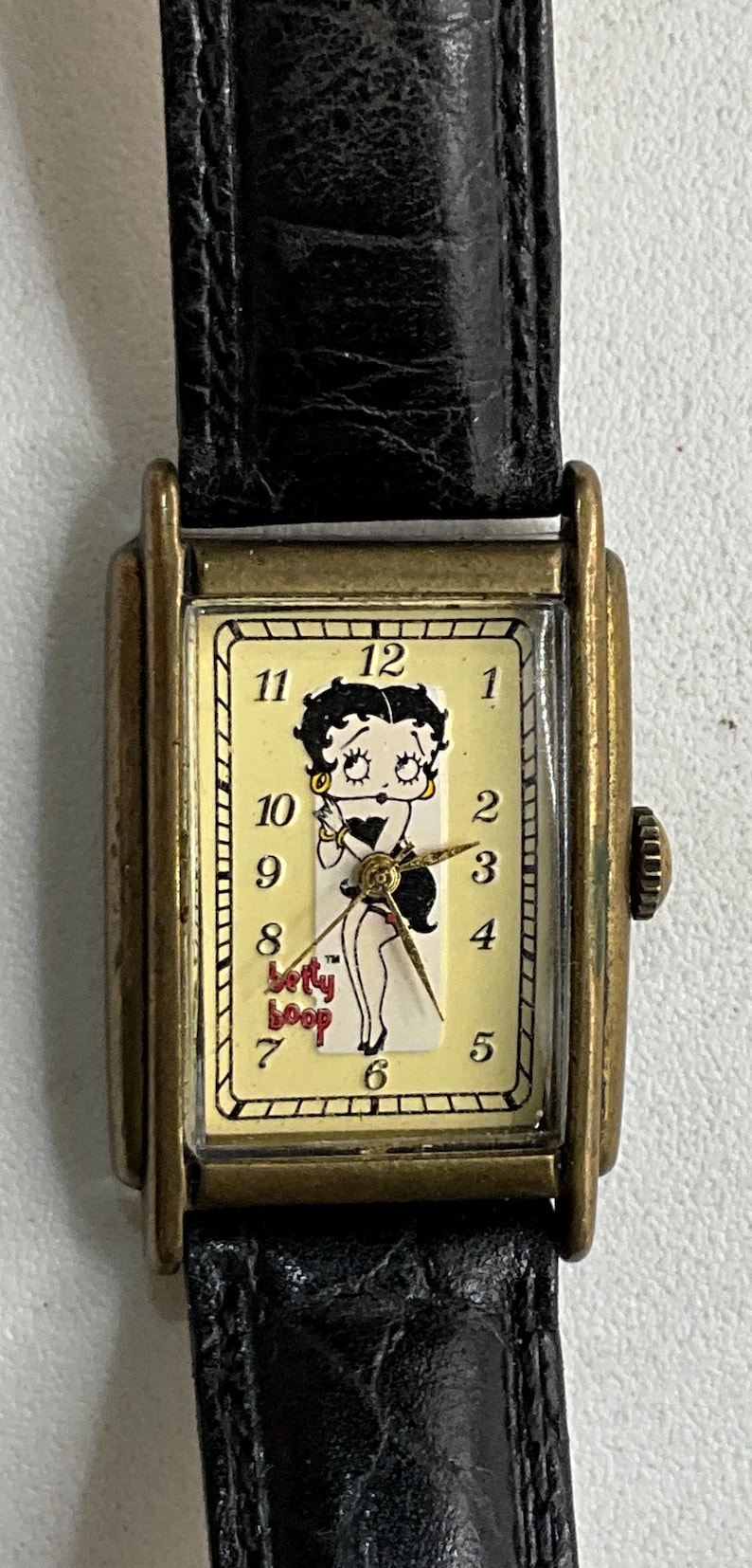 Peut inclure: Une montre-bracelet rectangulaire avec un bracelet en cuir noir et un bo&icirc;tier dor&eacute;. Le cadran est de couleur cr&egrave;me et repr&eacute;sente le personnage de dessin anim&eacute; Betty Boop. Les aiguilles sont dor&eacute;es et la montre a une petite couronne.