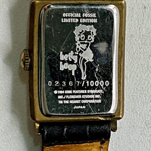 Peut inclure: Montre officielle Fossil en &eacute;dition limit&eacute;e avec Betty Boop. La montre a un bo&icirc;tier rectangulaire dor&eacute; avec un bracelet en cuir noir. Le cadran affiche le logo Betty Boop et le texte "0236 7/10000".