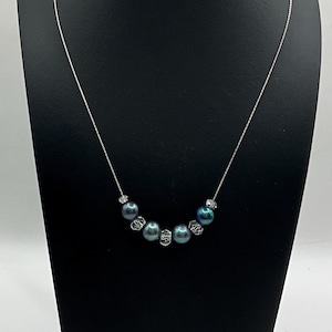 Puede incluir: Un delicado collar con perlas azul-verdosas oscuras intercaladas con pequeños acentos de cristal transparente. El collar se exhibe en un soporte de joyería negro, mostrando su elegante diseño y sutil brillo.