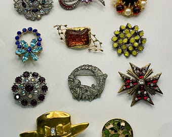 May include: Assortment of vintage brooches featuring various designs and colors, including a red and black floral design, a yellow and black geometric design, a blue and silver floral design, a silver and white floral design, a gold and green floral design, a gold and white floral design, a gold and red floral design, a gold and white floral design, and a gold and white floral design.
