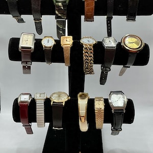 May include: A display of 17 wristwatches with various styles, colors, and materials. The watches are arranged on a black stand with multiple tiers. Some watches have gold, silver, or black bands, while others have leather or metal bands. The watches are a mix of round, square, and rectangular shapes.
