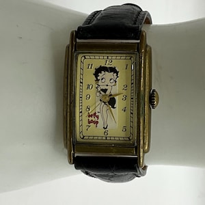 Peut inclure: Montre-bracelet rectangulaire vintage avec un bracelet en cuir noir et un bo&icirc;tier dor&eacute;. Le cadran de la montre pr&eacute;sente un dessin de Betty Boop et les mots "Betty Boop". La montre a un design classique.