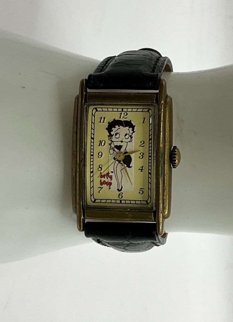 Peut inclure: Montre-bracelet rectangulaire vintage avec un bracelet en cuir noir et un bo&icirc;tier dor&eacute;. Le cadran de la montre pr&eacute;sente un dessin de Betty Boop et les mots "Betty Boop". La montre a un design classique.