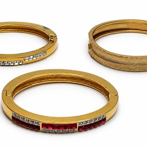 Op de afbeelding: Drie goudkleurige scharnierende armbanden. Eén is voorzien van doorzichtige stenen, één heeft een gestructureerd kruispatroon en de derde toont een rij afwisselend rode en doorzichtige stenen. Elke armband is cirkelvormig met een sluiting.