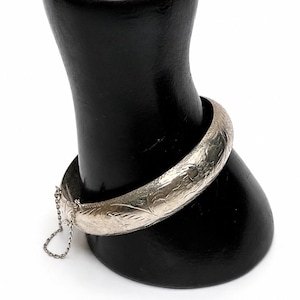 May include: A silver-toned hinged bangle bracelet featuring an intricate engraved floral pattern across its surface. The bracelet includes a secure clasp mechanism and a delicate attached safety chain, providing a functional and decorative design.