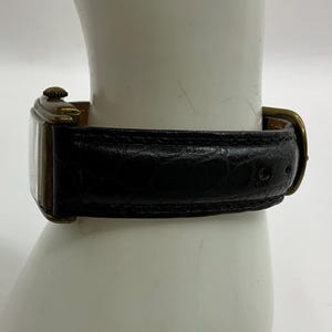 Peut inclure: Une montre-bracelet rectangulaire vintage avec un bracelet en cuir noir et un bo&icirc;tier dor&eacute;. La montre est attach&eacute;e avec une boucle dor&eacute;e. La montre est pr&eacute;sent&eacute;e sur une surface blanche.