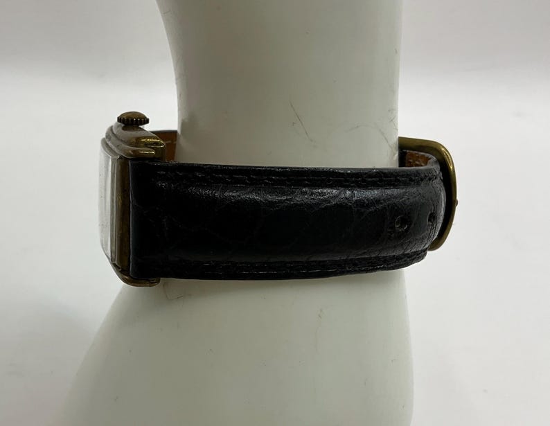 Peut inclure: Une montre-bracelet rectangulaire vintage avec un bracelet en cuir noir et un bo&icirc;tier dor&eacute;. La montre est attach&eacute;e avec une boucle dor&eacute;e. La montre est pr&eacute;sent&eacute;e sur une surface blanche.