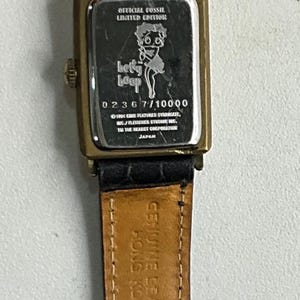 Peut inclure: Une montre-bracelet Fossil Betty Boop en &eacute;dition limit&eacute;e. La montre a un bo&icirc;tier rectangulaire dor&eacute; avec un bracelet en cuir noir. Le cadran de la montre pr&eacute;sente un graphisme Betty Boop et le texte "Betty Boop".