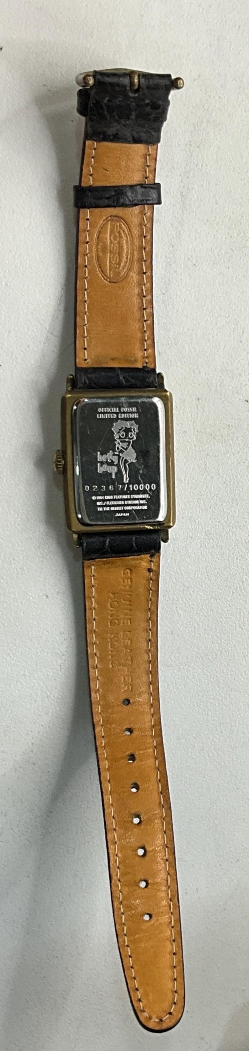 Peut inclure: Une montre-bracelet Fossil Betty Boop en &eacute;dition limit&eacute;e. La montre a un bo&icirc;tier rectangulaire dor&eacute; avec un bracelet en cuir noir. Le cadran de la montre pr&eacute;sente un graphisme Betty Boop et le texte "Betty Boop".