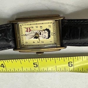 Peut inclure: Une montre-bracelet vintage avec un bo&icirc;tier rectangulaire dor&eacute; et un bracelet en cuir noir. Le cadran de la montre pr&eacute;sente un personnage de dessin anim&eacute;. Le bracelet mesure environ 23 cm de long.