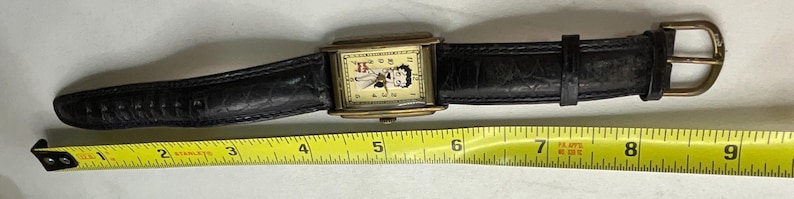 Peut inclure: Une montre-bracelet vintage avec un bo&icirc;tier rectangulaire dor&eacute; et un bracelet en cuir noir. Le cadran de la montre pr&eacute;sente un personnage de dessin anim&eacute;. Le bracelet mesure environ 23 cm de long.