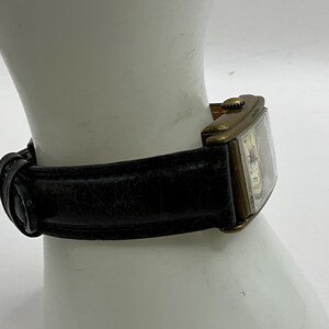 Peut inclure: Une montre-bracelet rectangulaire vintage avec un bo&icirc;tier dor&eacute; et un bracelet en cuir noir. Le cadran de la montre est visible, avec un cadran et des aiguilles de couleur claire. Le bracelet est fix&eacute; avec une boucle.