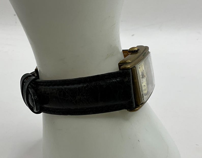 Peut inclure: Une montre-bracelet rectangulaire vintage avec un bo&icirc;tier dor&eacute; et un bracelet en cuir noir. Le cadran de la montre est visible, avec un cadran et des aiguilles de couleur claire. Le bracelet est fix&eacute; avec une boucle.