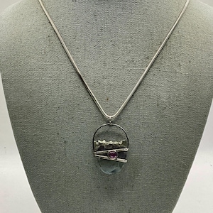 May include: A silver-toned snake chain necklace featuring a pendant with a smooth, dark, rounded stone. The stone is accented by a small, round, magenta-colored gemstone set within a silver wire frame that wraps around the center of the stone.