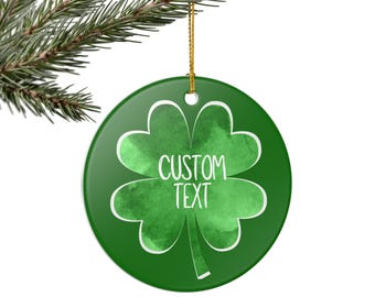 Peut inclure: Ornement rond en céramique verte avec un motif de trèfle blanc et l'inscription "CUSTOM TEXT". L'ornement est suspendu par une ficelle dorée et présenté sur une branche de pin.