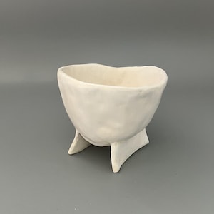 May include: A handmade, off-white ceramic bowl with a rounded body and three angular legs. The bowl has a smooth, matte finish and a slightly irregular rim. This unique piece is perfect for home decor.