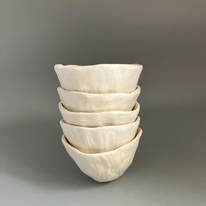 May include: A stack of six small, off-white ceramic bowls. Each bowl has a textured surface and a slightly irregular, wavy rim. The bowls are stacked neatly, with the bottom bowl resting on a gray surface.