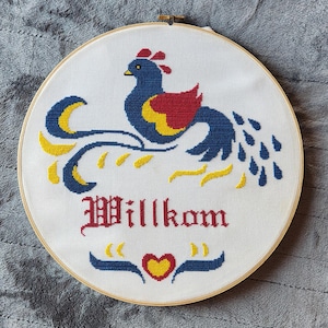 May include: Cross-stitch embroidery in a wooden hoop. The design features a blue bird with red and yellow accents, and the word "Willkom" in red gothic lettering. A blue banner with a yellow heart is below.