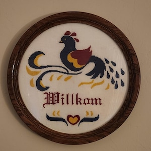 May include: A round, framed cross-stitch featuring a stylized bird with a long tail, in shades of blue, yellow, and burgundy. The word "Willkom" is stitched below the bird, with a heart and decorative elements. The frame is made of dark wood.
