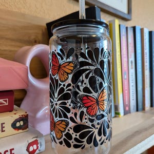May include: A clear glass tumbler with a black lid and straw. The tumbler is decorated with orange and black monarch butterfly designs and black floral patterns. The tumbler is sitting on a wooden shelf.