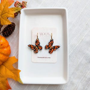 May include: A pair of butterfly-shaped earrings featuring orange and black wings with white accents, attached to gold-tone hoops. The earrings are displayed on a rectangular card labeled Moon & Cats with the website Moonandcats.com printed below.
