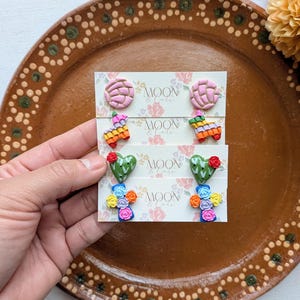 Op de afbeelding: Vier paar oorknopjes op witte displaykaarten met het label Moon & Cats. De ontwerpen omvatten roze concha-broodjes, veelkleurige piñata's, groene hartvormige cactussen met rode bloemen en blauwe kruisjes versierd met kleine gele, roze en blauwe roosjes.