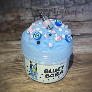 Könnte beinhalten: Ein hellblaues Schleimgefäß mit Deckel, verziert mit Bluey-Charms und einem Miniatur-Boba-Getränk. Der Behälter hat ein Etikett mit der Aufschrift "BLUEY BOBA" und einer Bluey-Charakterillustration. Der Hintergrund ist eine Holzoberfläche.
