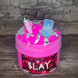 Könnte beinhalten: Rosa Schleim mit silbernem Glitzer in einem durchsichtigen Glas mit einem schwarz-weißen Etikett, auf dem "SLAY" steht. Das Glas hat eine rosa-weiße Mini-Wasserflasche und ein weißes Telefon mit einem schwarzen Bildschirm, auf dem "08:54" steht, auf dem Schleim.