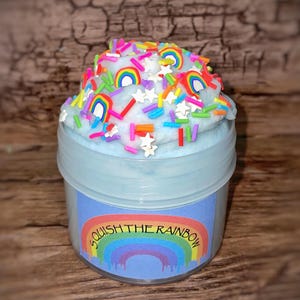 Könnte beinhalten: Ein durchsichtiges Plastikglas gefüllt mit hellblauem Schleim, der mit bunten Regenbogenstreuseln und weißen Sternen bedeckt ist. Das Glas hat ein blaues Etikett mit der Aufschrift "Squish the Rainbow".