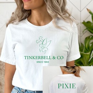 May include: White t-shirt with a green line drawing of a fairy and flowers. The text "TINKERBELL & CO SINCE 1904" is printed below the image. The back of the shirt has the word "PIXIE" printed in green.