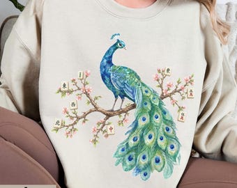 May include: A sand-colored crewneck sweatshirt featuring a graphic illustration of a blue and green peacock perched on a flowering branch. The design includes several mahjong tiles with Chinese characters integrated into the floral branches.