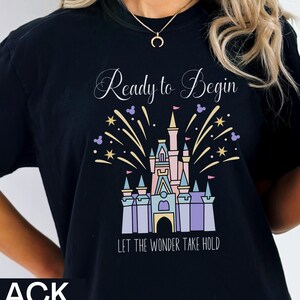 K&ouml;nnte beinhalten: Schwarzes T-Shirt mit einem wei&szlig;en Aufdruck von Cinderellas Schloss mit Feuerwerk und dem Text "Ready to Begin" und "Let the Wonder Take Hold".