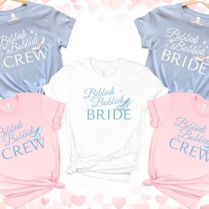 May include: Set of five t-shirts with a light blue and pink color scheme. The shirts feature the text "Bibbidi Bobbidi" with a Cinderella glass slipper graphic. Two shirts say "Crew" and two shirts say "Bride".