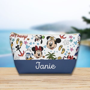 May include: A white and blue toiletry bag with a Disney theme. The bag features Mickey Mouse, Minnie Mouse, Donald Duck, and Goofy. The bag has a blue bottom with the name "Janie" written in white.