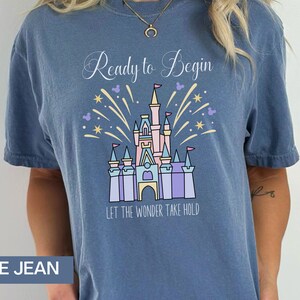 K&ouml;nnte beinhalten: Ein T-Shirt aus Jeansblau mit einer wei&szlig;en Grafik von Cinderellas Schloss mit Feuerwerk und dem Text "Ready to Begin" und "Let the Wonder Take Hold".