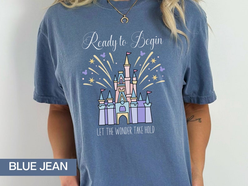 K&ouml;nnte beinhalten: Ein T-Shirt aus Jeansblau mit einer wei&szlig;en Grafik von Cinderellas Schloss mit Feuerwerk und dem Text "Ready to Begin" und "Let the Wonder Take Hold".