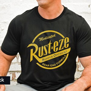 May include: Black crew neck t-shirt with a yellow and gold graphic. The graphic reads "Rust-eze Bumper Ointment Rear End Formula" in a circular design. The shirt is made of a soft material.