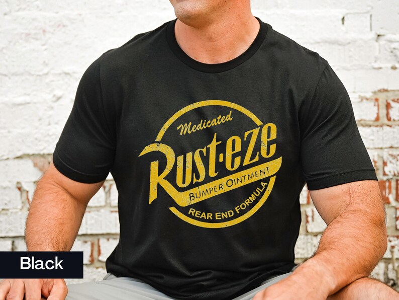 May include: Black crew neck t-shirt with a yellow and gold graphic. The graphic reads "Rust-eze Bumper Ointment Rear End Formula" in a circular design. The shirt is made of a soft material.