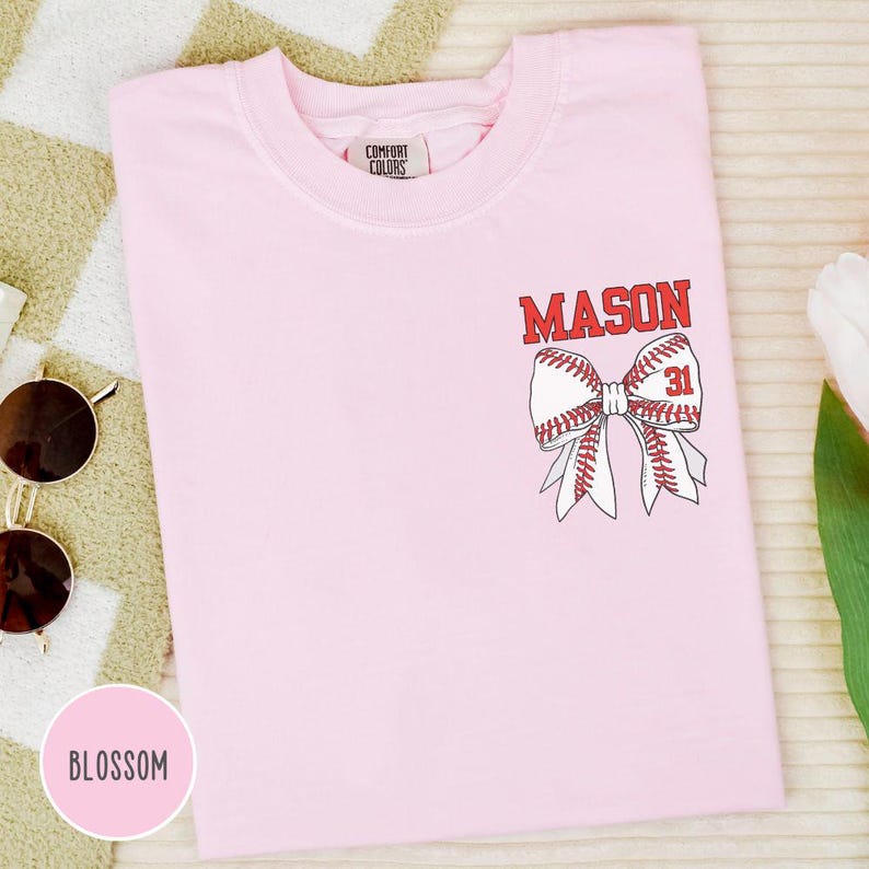 May include: A light pink t-shirt with a baseball bow design. The bow is made of baseballs with red stitching and the name "MASON" in red letters above it. The number "31" is inside the bow. The shirt has a crew neck and short sleeves.